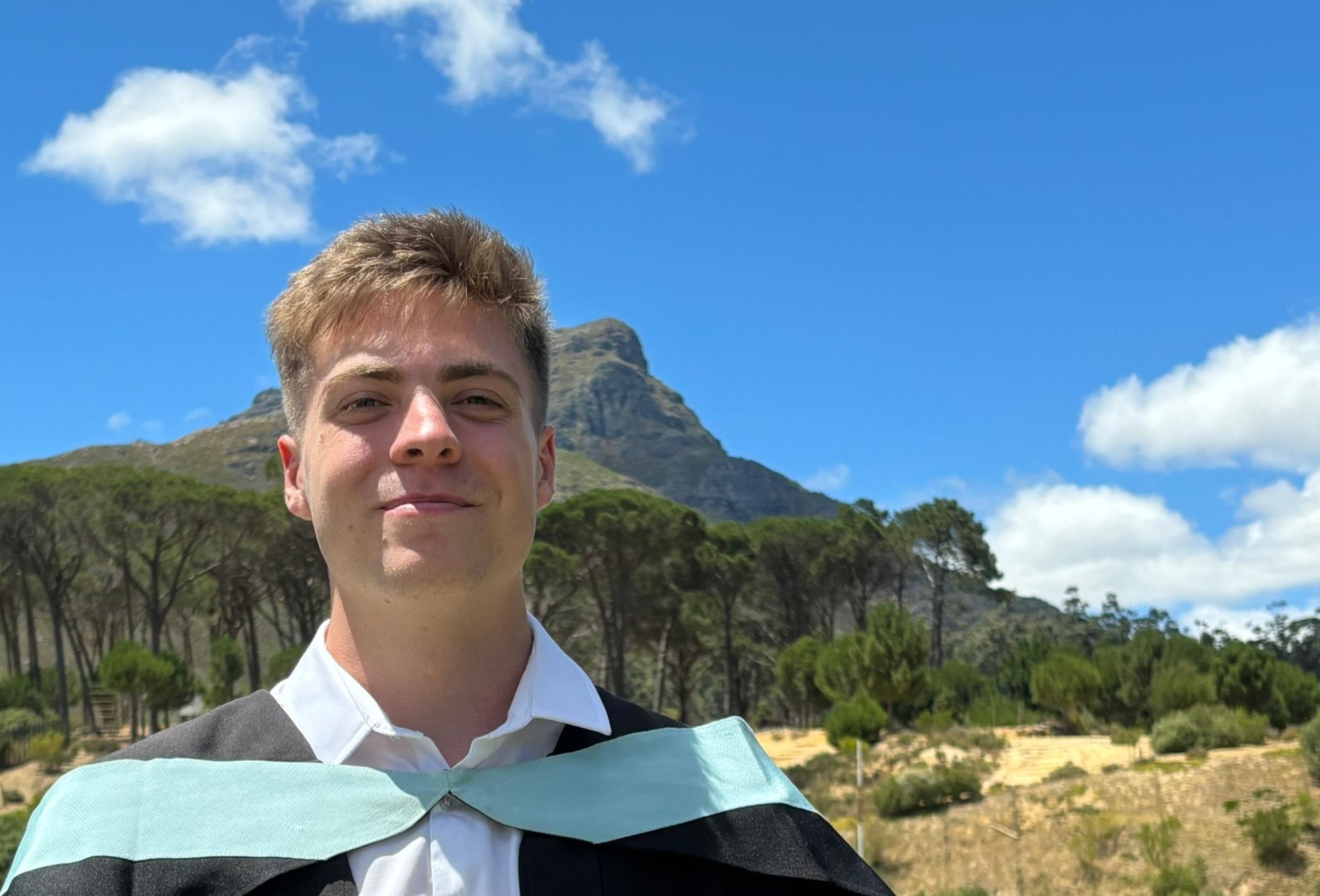 Mikael Pretorius outdoors, a mountain and pine trees behind him under a blue sky